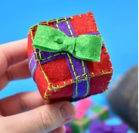 Homemade catnip present. Red box with purple ribbon and a green bow on top being held in a person's hand with a bright blue background
