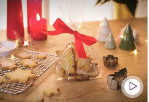 Star shaped biscuits in a plastic bag on a wooden table with a red bow tied around the top of the bag. Baking wire rack on the table too with star shaped biscuits cooling on top